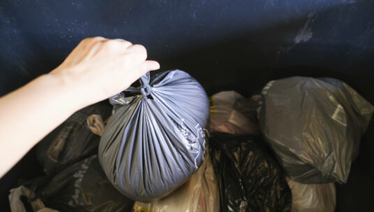 Kuvassa näkyy roska-astian sisäpuoli ja käsi, joka on pudottamassa roskapussia astiaan.